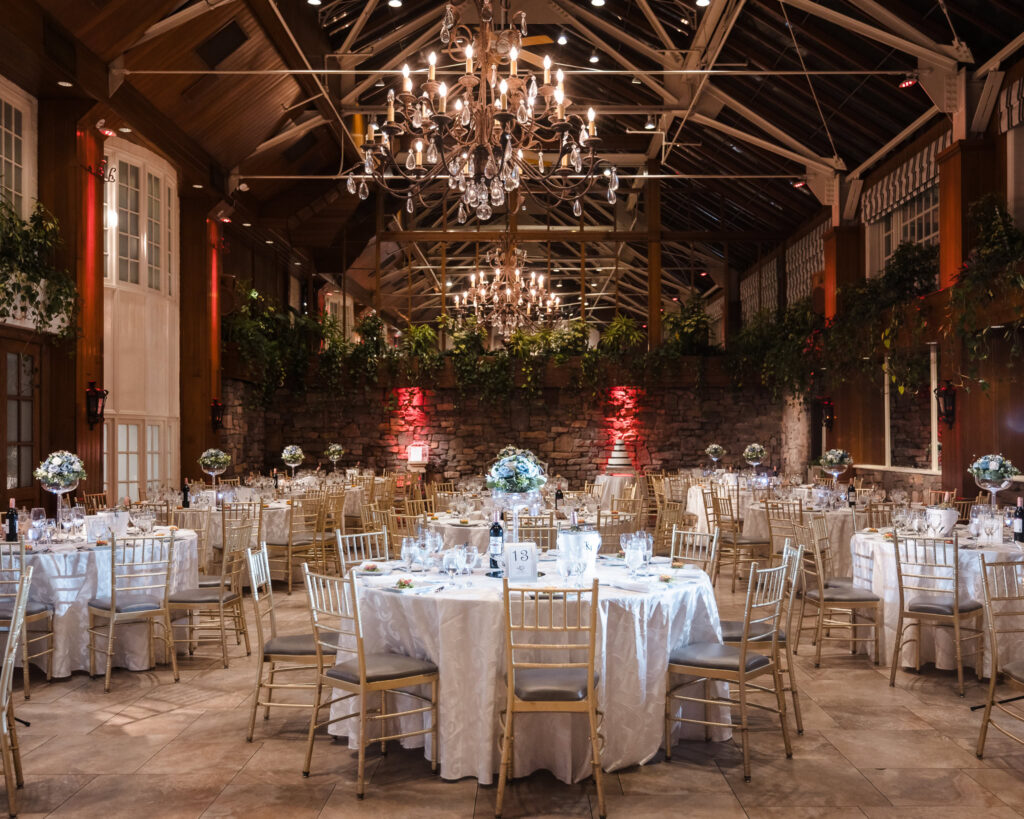 Elegant reception setup at Fox Hollow’s Winter Garden with candlelit tables, gold chairs, floral centerpieces, and chandeliers under a vaulted glass ceiling.