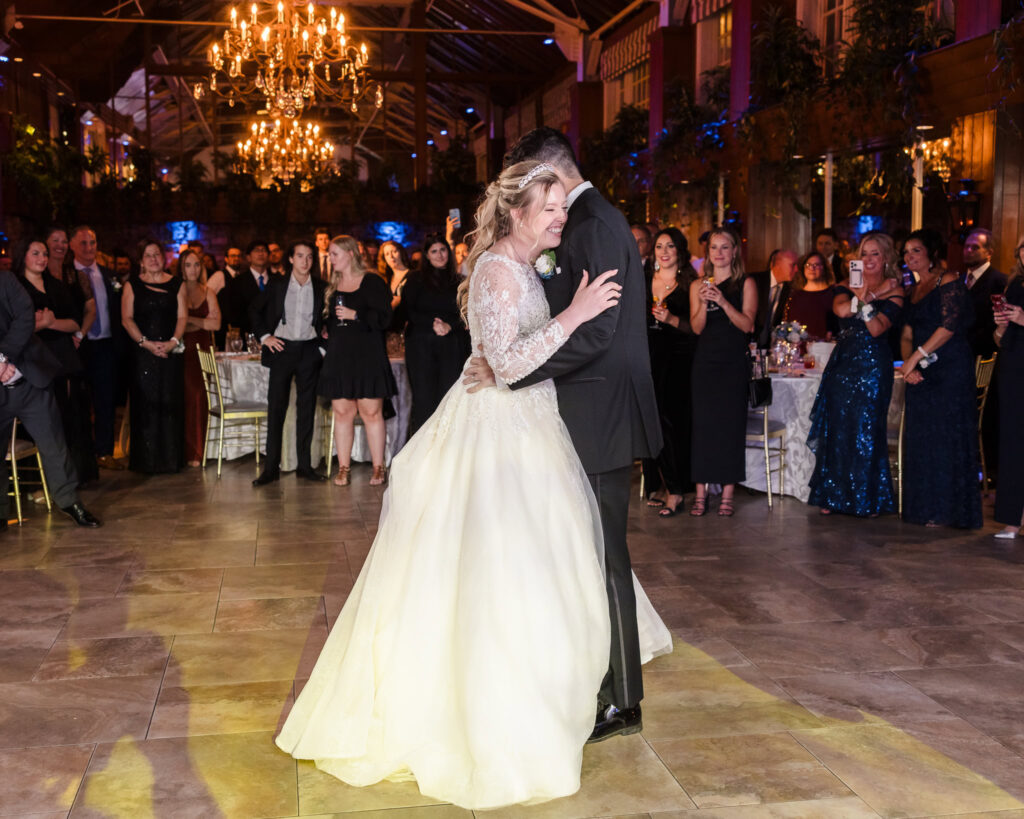 Bride and groom share their first dance surrounded by guests in the warmly lit Winter Garden at Fox Hollow.