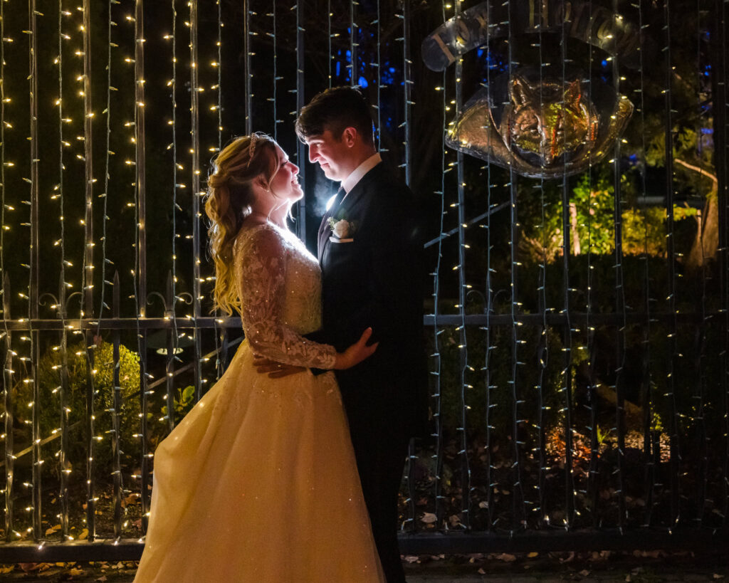 Bride and groom share an intimate nighttime moment beneath twinkling string lights at the Fox Hollow gate, softly illuminated as they gaze at each other