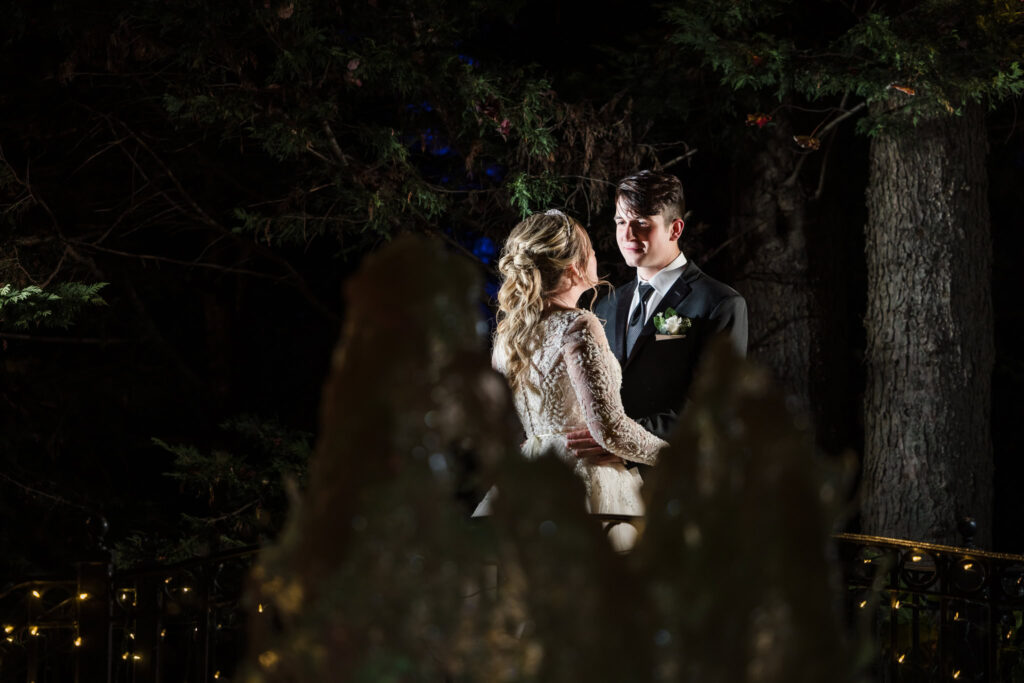 Bride and groom share an illuminated evening portrait in the woods at Fox Hollow, standing closely together near soft bokeh-lit trees.