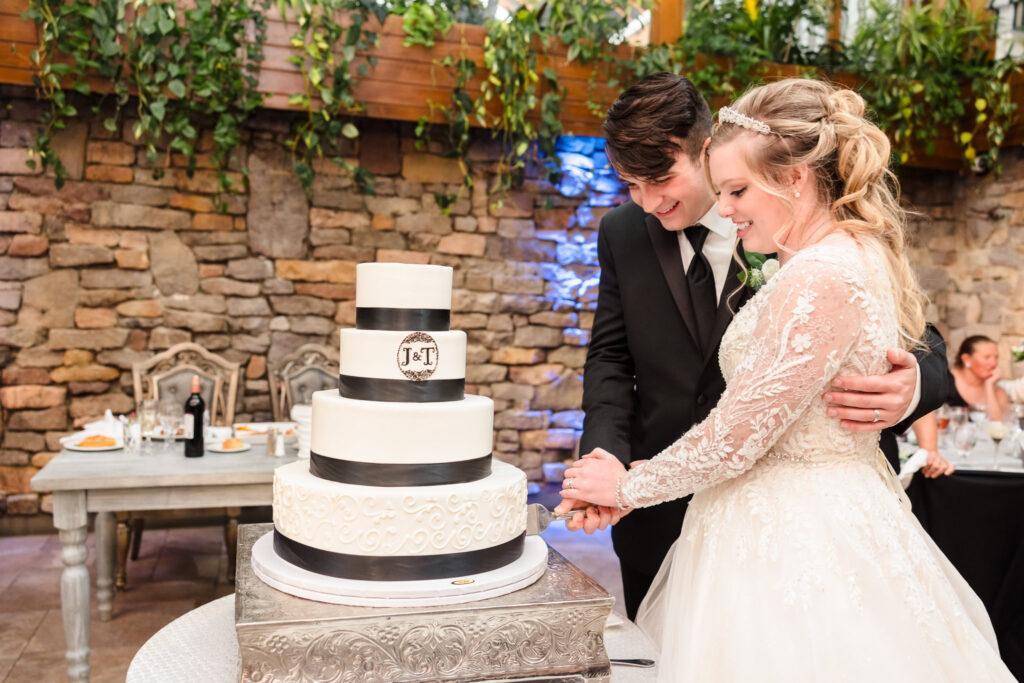 Bride and groom cut their tiered white wedding cake together, sharing a sweet moment in front of a stone wall.