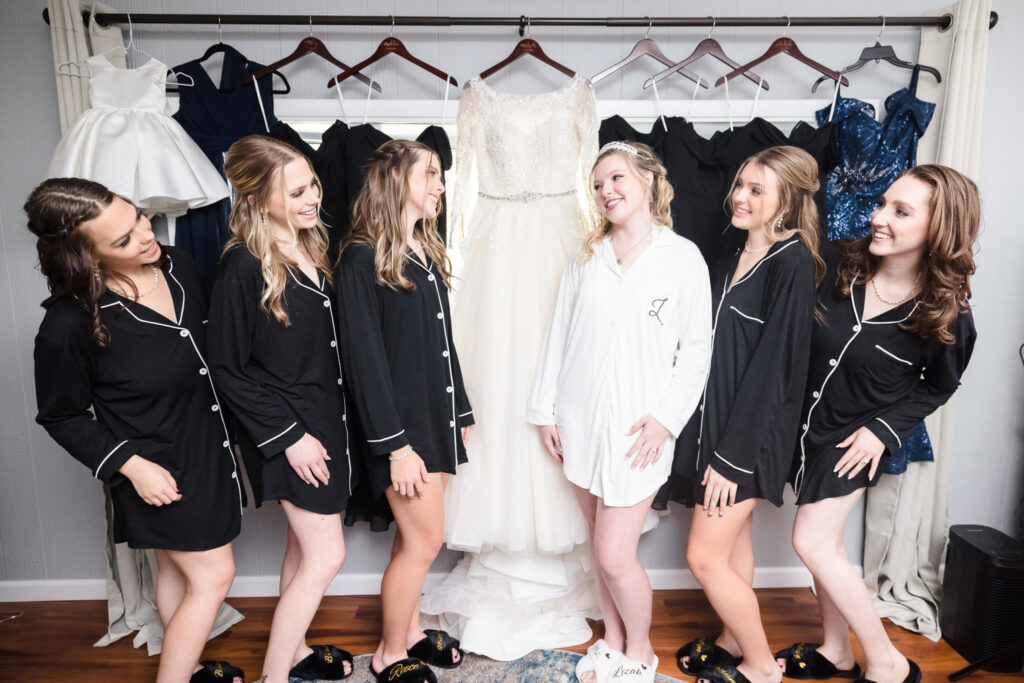 Bride and bridesmaids in matching pajamas smile and laugh together in front of the wedding gown and dresses during a getting ready moment at home.