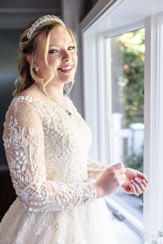 Bride smiles softly while getting ready at home, standing near a window in her embroidered lace wedding gown.