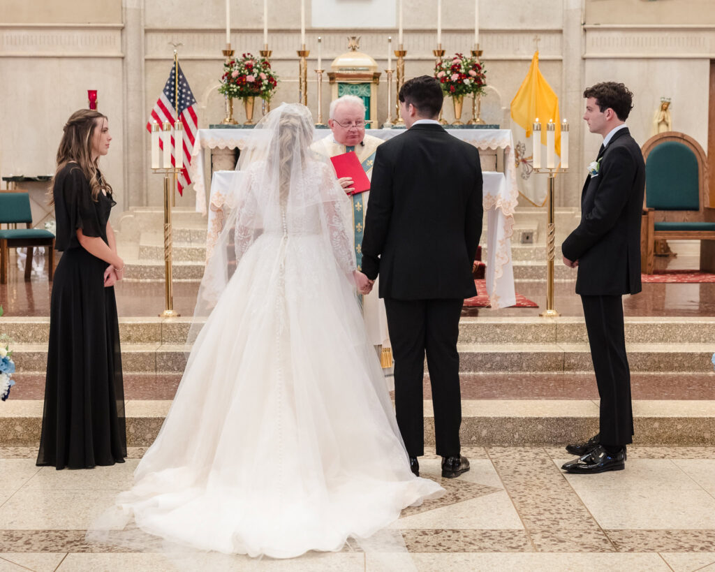 Bride and groom stand hand-in-hand before the altar at St. Patrick’s Church during their wedding ceremony, flanked by their attendants.