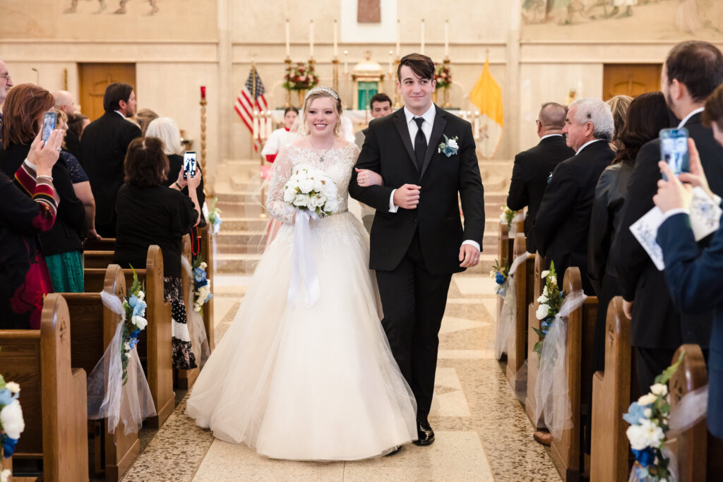 Bride and groom walk down the aisle of St. Patrick’s Church in Huntington, NY, smiling joyfully as guests capture the moment.