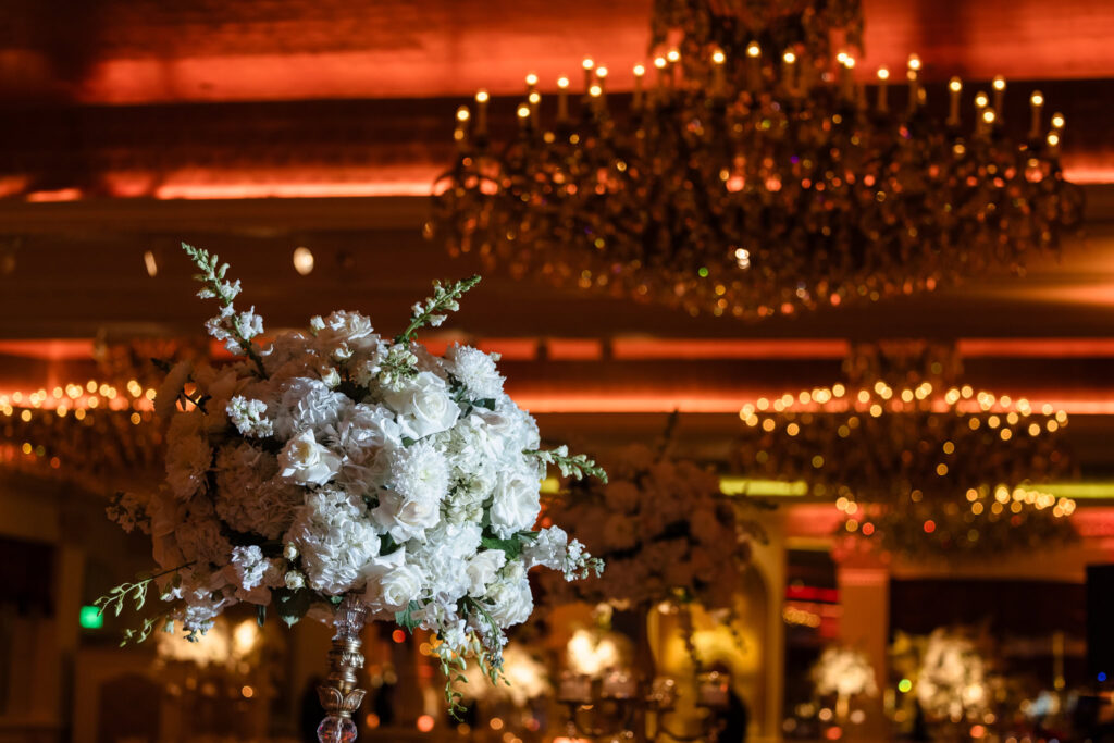 A luxurious white floral centerpiece glows under the warm amber lighting and ornate crystal chandeliers of the Grand Ballroom at Garden City Hotel—highlighting the romantic ambiance of an upscale Long Island wedding reception.

