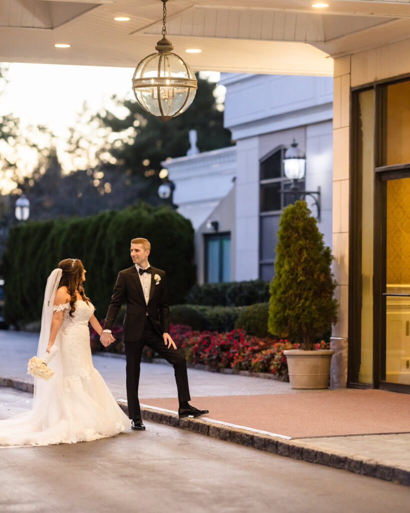 Bride and groom share a joyful moment outside the entrance of the Garden City Hotel in Garden City, NY, surrounded by manicured hedges, blooming florals, and warm architectural lighting—capturing the timeless charm of a Long Island wedding venue.

