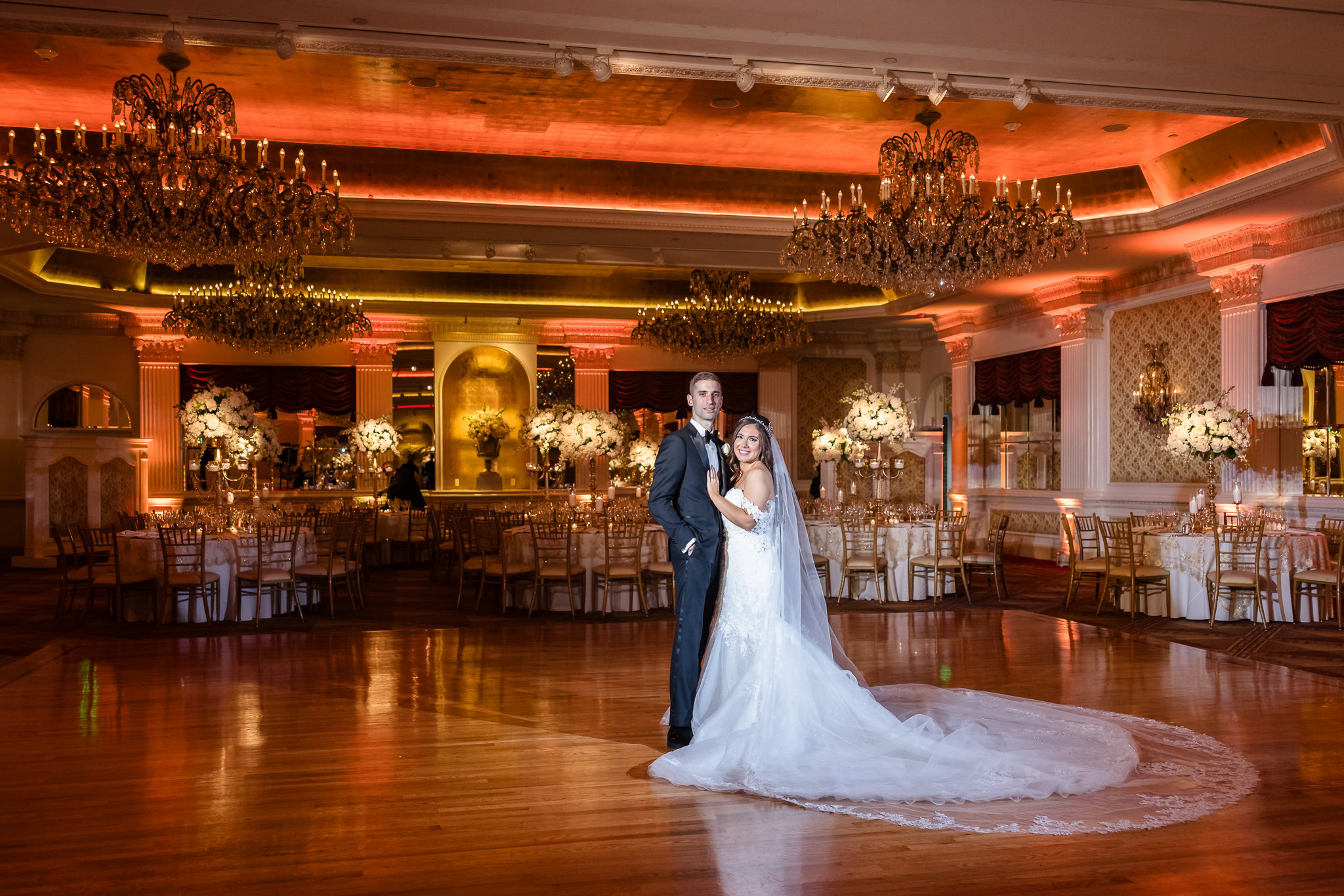 A bride and groom pose elegantly in the Grand Ballroom of the Garden City Hotel, surrounded by lavish floral centerpieces, gold chiavari chairs, and grand chandeliers glowing with amber uplighting—capturing timeless elegance on Long Island.