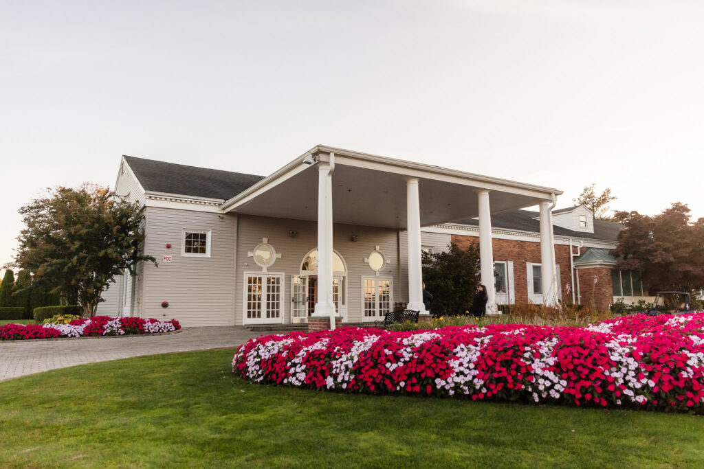 The front entrance of Glen Head Country Club, featuring grand white columns, manicured lawns, and vibrant pink and white flowers in full bloom during golden hour.