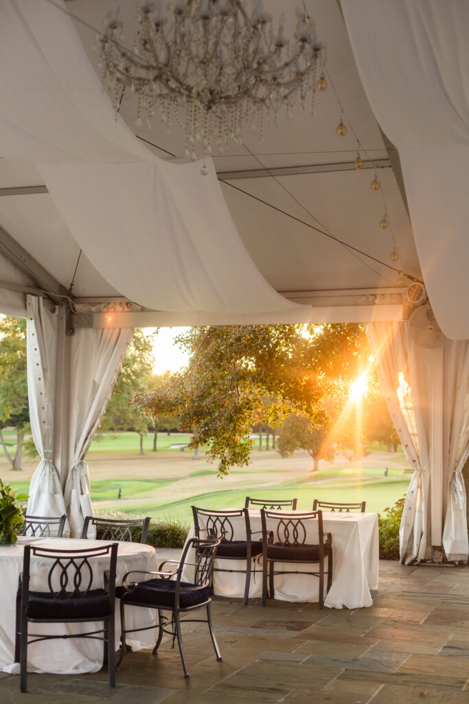Sunset glow filters through white drapery on the Clubhouse Terrace at Glen Head Country Club, with elegant black chairs and white linens set against a backdrop of rolling green lawns.