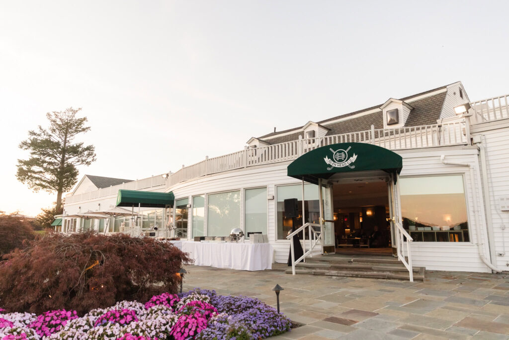 The back patio of Glen Head Country Club at sunset, featuring white-paneled architecture, green awnings with the club emblem, slate stone flooring, and vibrant flower beds in pinks and purples.