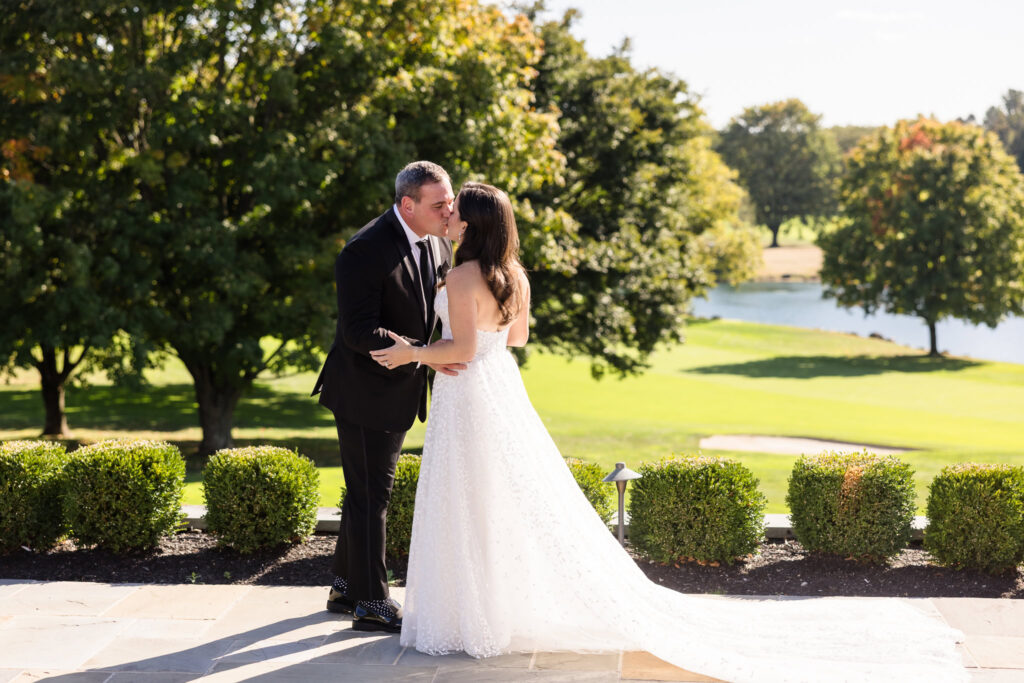 A bride in a flowing lace gown kisses her groom on the terrace, framed by manicured hedges, lush trees, and a sparkling pond in the background.