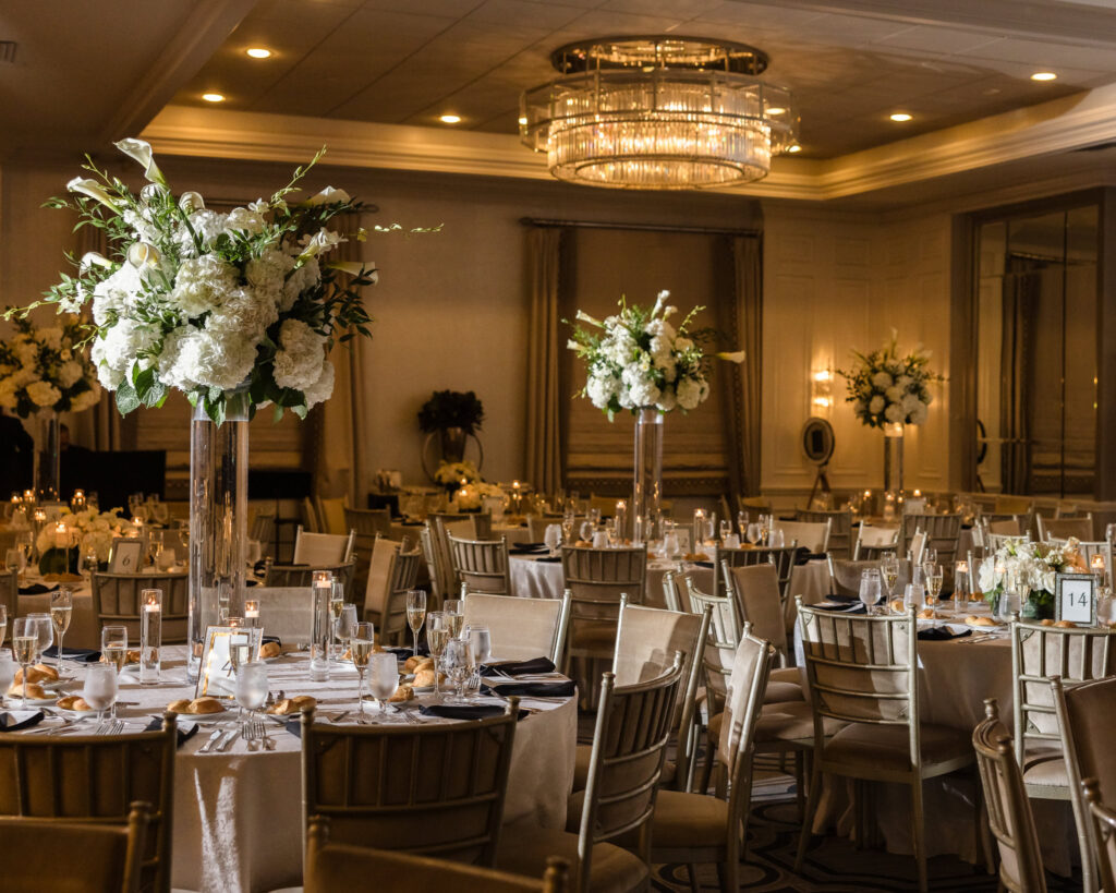 An elegant wedding reception setup inside the Grand Ballroom at Glen Head Country Club, featuring tall floral centerpieces, gold chairs, soft candlelight, and a modern crystal chandelier overhead.