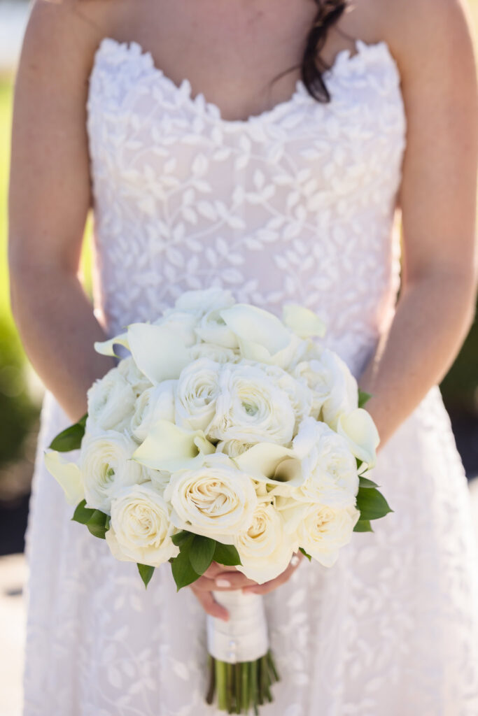 Close-up of a bride holding an elegant bouquet of cream roses and calla lilies, with her lace-embroidered gown softly visible in the background.
