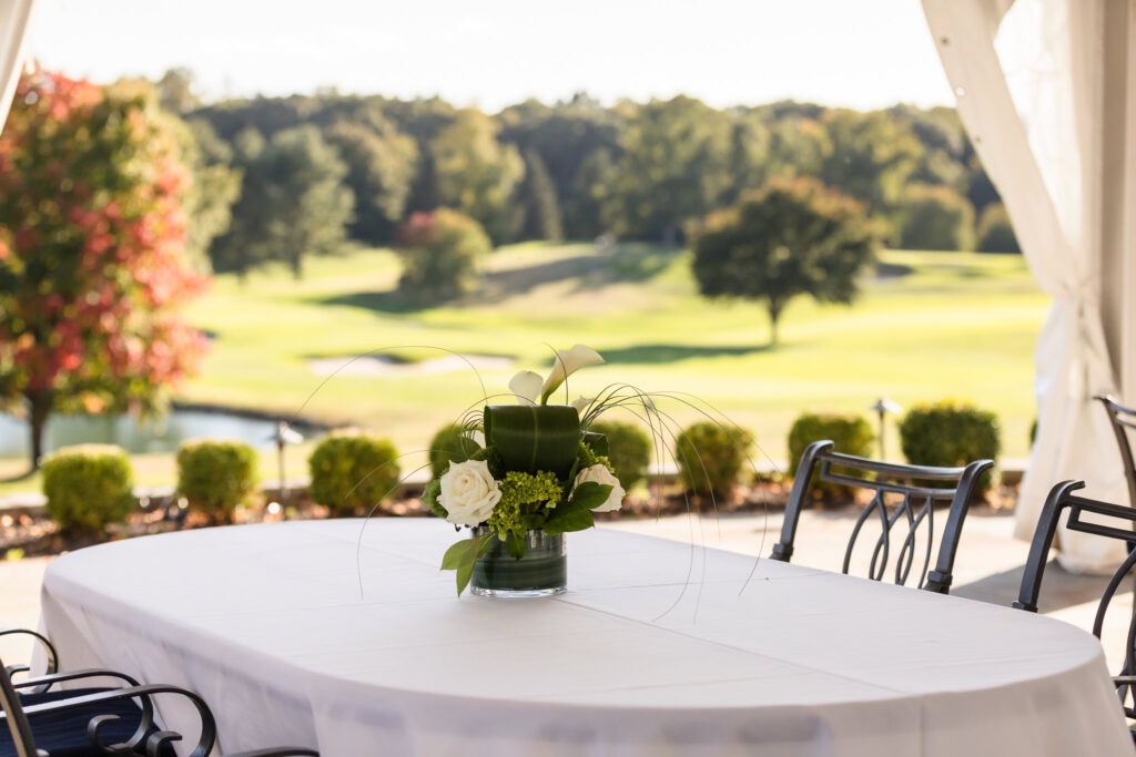 A refined white centerpiece featuring roses and calla lilies sits atop a linen-covered table on the Clubhouse Terrace, with sweeping views of the golf course and early autumn foliage in the background.