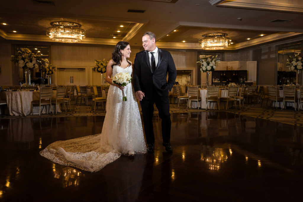 Bride and groom walk hand-in-hand across the polished dance floor of the Grand Ballroom, surrounded by elegant reception décor.