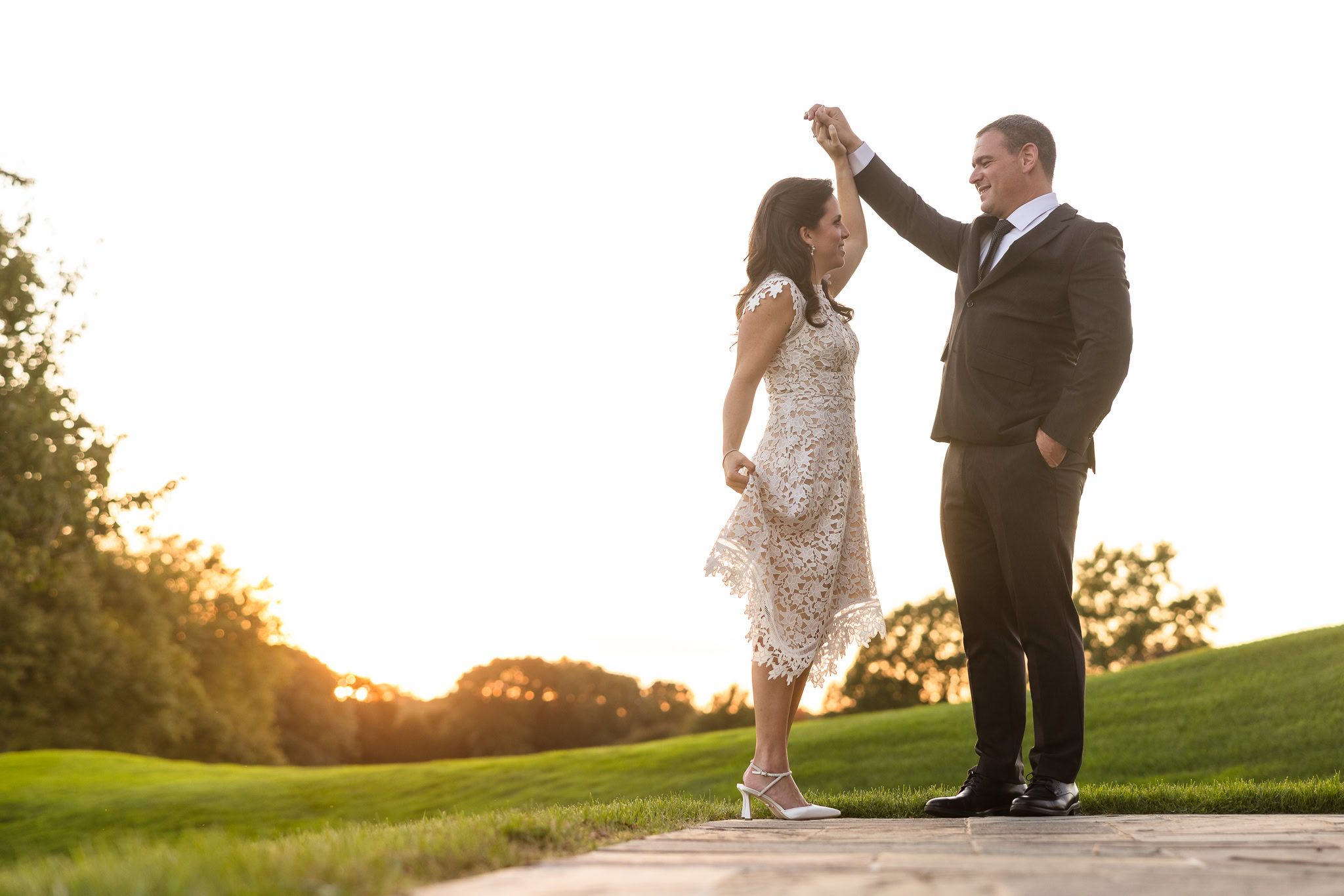 Groom twirls his bride at golden hour on the terrace of Glen Head Country Club, with rolling green hills and a glowing sunset in the background.
