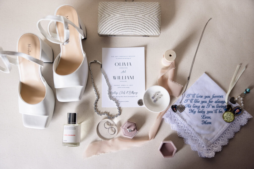 Bridal flat lay featuring white heels, wedding invitation, pearl clutch, perfume, jewelry, and embroidered handkerchief for Olivia and William's Heritage Club wedding.