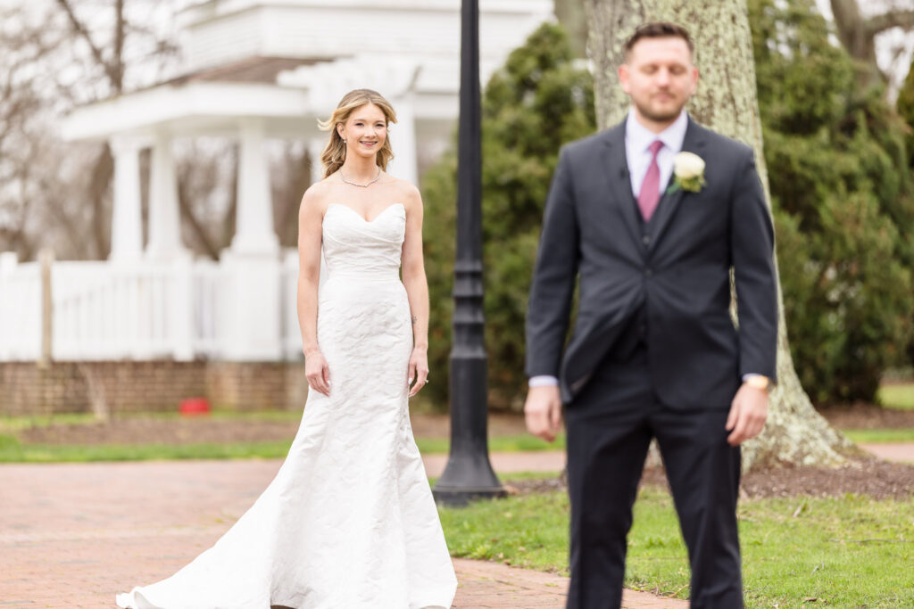 Bride and groom standing apart but in focus near The Heritage Club gardens, framed by manicured trees and spring greenery.