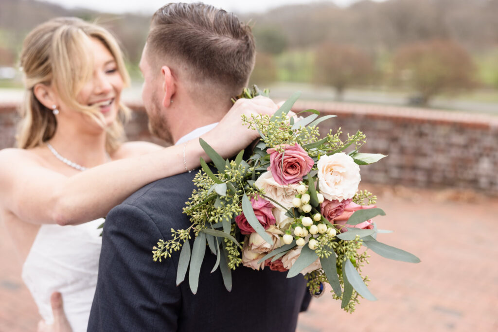 Close-up of bride and groom embracing outdoors, with the bride’s romantic spring bouquet of roses and greenery in sharp focus.