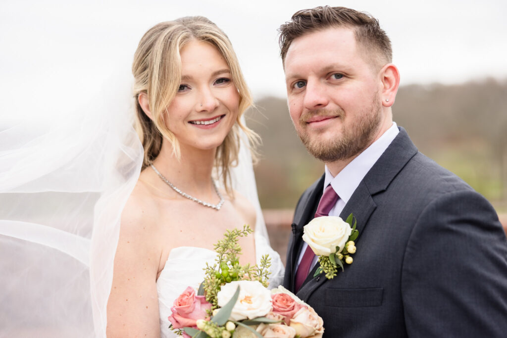 Bride and groom smiling together with soft veil movement and a blush-toned bouquet, captured outdoors in natural light.