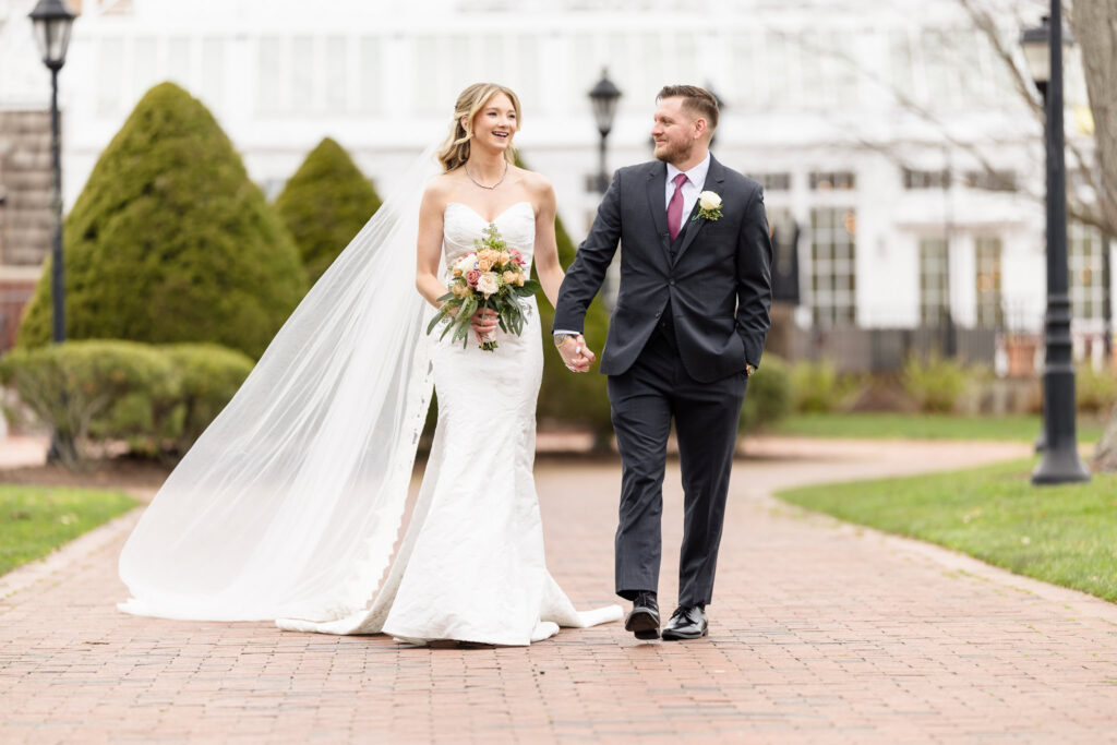 Bride and groom holding hands and smiling while walking along a brick path outside The Heritage Club, with the bride’s veil flowing in the spring breeze.