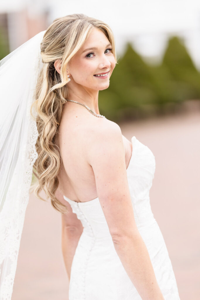 Close-up of bride glancing over her shoulder in natural light, with soft curls, a veil, and a serene spring backdrop.