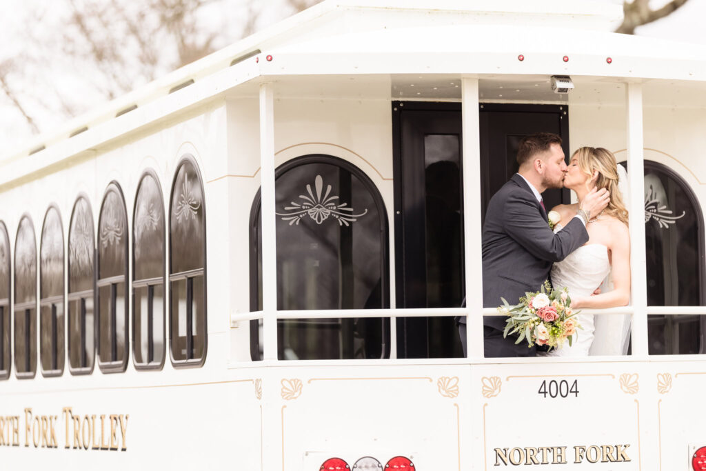 Bride and groom sharing a kiss aboard a classic white North Fork Trolley, offering a charming and joyful transportation moment.