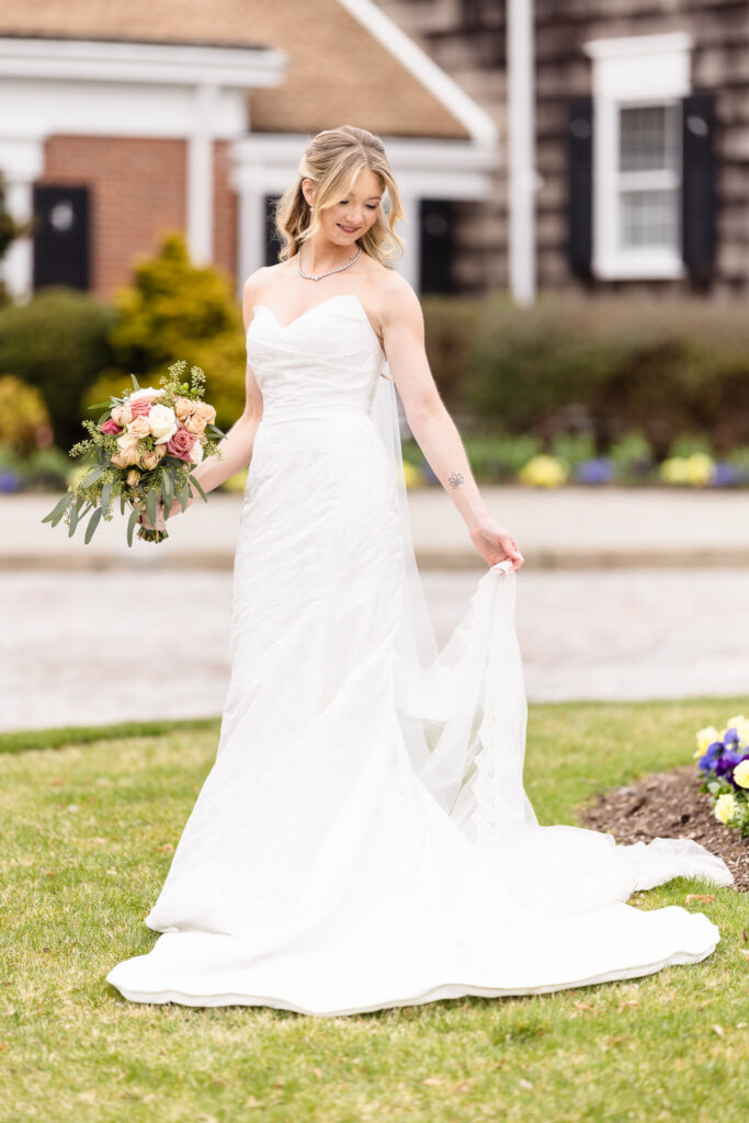 Full-length bridal portrait of bride lifting her dress slightly and holding a bouquet, standing on the grass outside The Heritage Club.