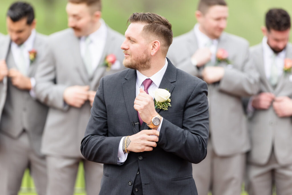 Groom in a dark suit with burgundy tie adjusting his cufflink while smiling, with groomsmen blurred in the background.