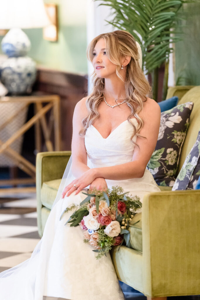 Bride seated on a green velvet chair with her bouquet in hand, framed by lush decor and soft light at The Heritage Club.