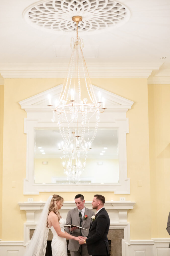 Bride and groom holding hands in front of their officiant during their indoor wedding ceremony at The Heritage Club, beneath an elegant chandelier.
