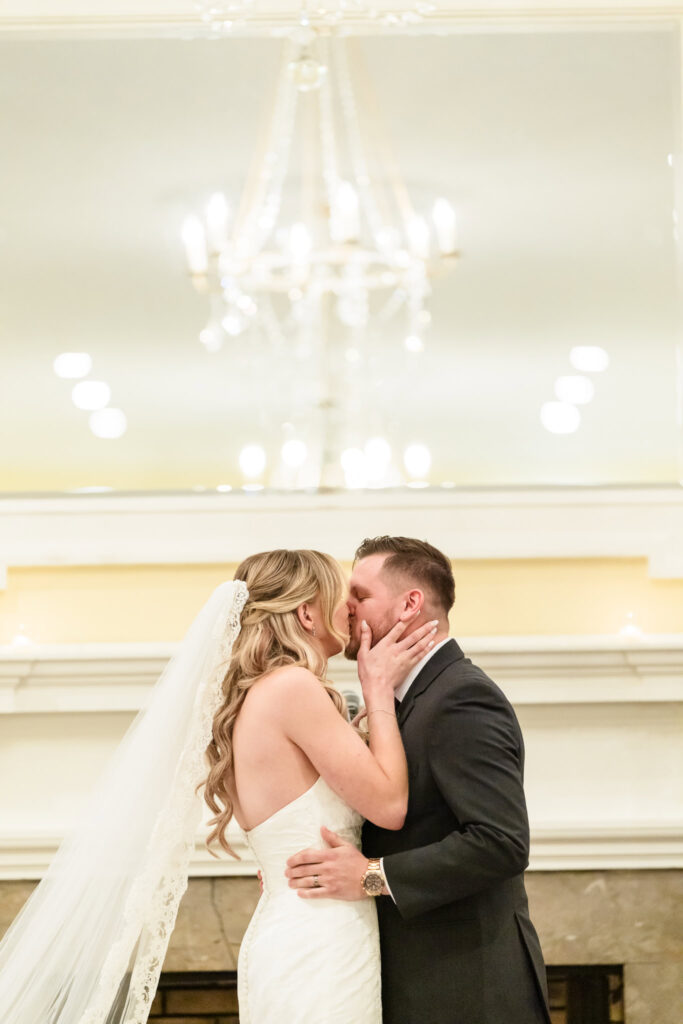 Bride and groom share their first kiss in a warmly lit ceremony space, with a chandelier glowing softly overhead.