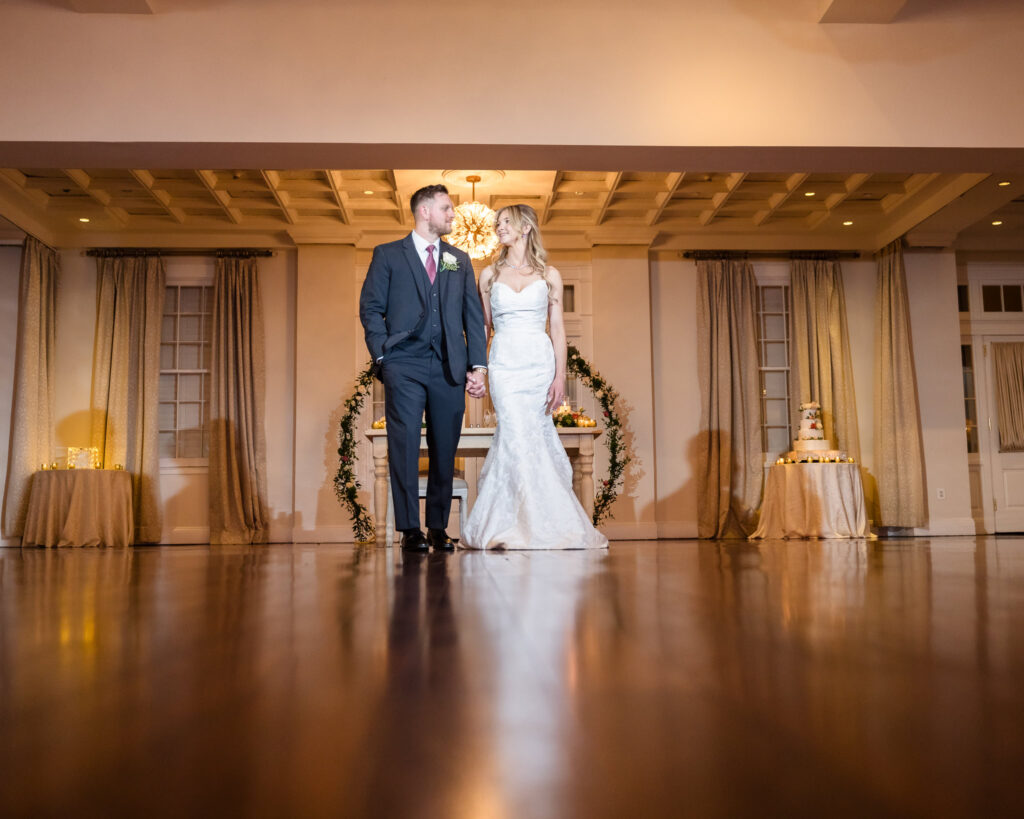 Bride and groom holding hands, gazing at each other on the center of the ballroom floor with cake and sweetheart table in the background, bathed in soft ambient light.