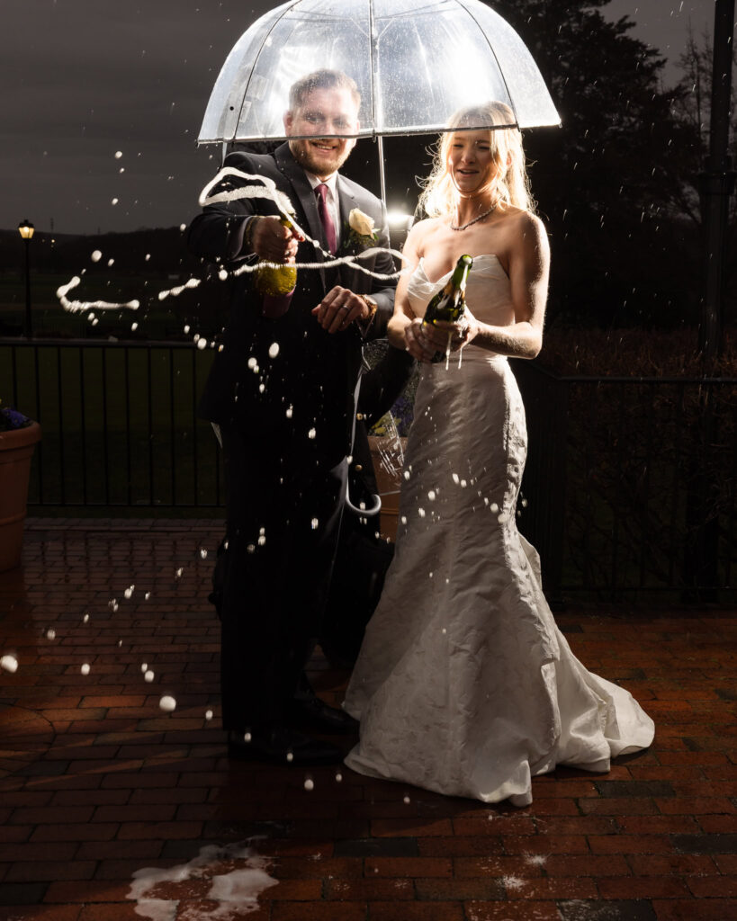Bride and groom laughing under a clear umbrella as champagne sprays in celebration during a nighttime portrait at The Heritage Club.