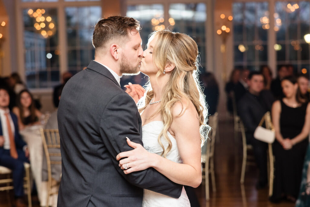 Bride and groom share a kiss during their first dance in a warmly lit reception room, surrounded by seated guests and sparkling bokeh lights.