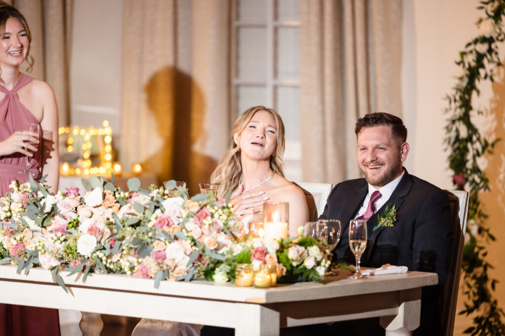 Bride and groom seated at a floral-adorned sweetheart table, smiling and laughing as a bridesmaid delivers a toast during their Heritage Club wedding reception.