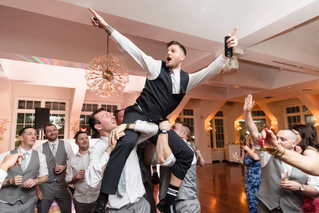 Groomsman raised by friends during a high-energy wedding reception dance at The Heritage Club, celebrating with arms outstretched.