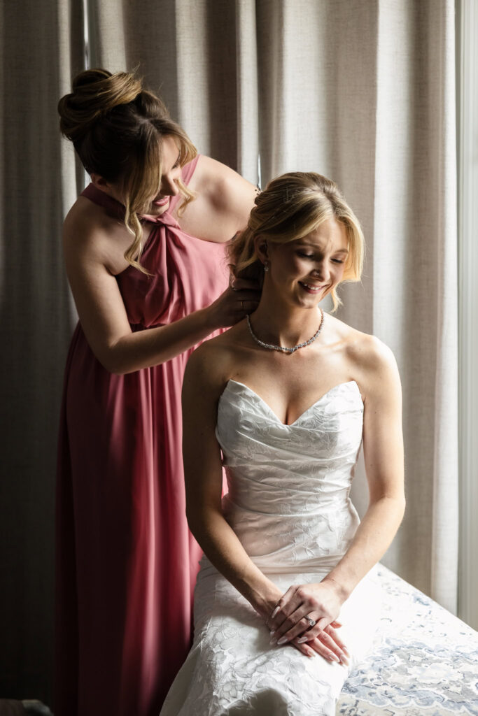Bridesmaid helps bride put on a necklace in a softly lit room, highlighting a calm, emotional getting-ready moment.
