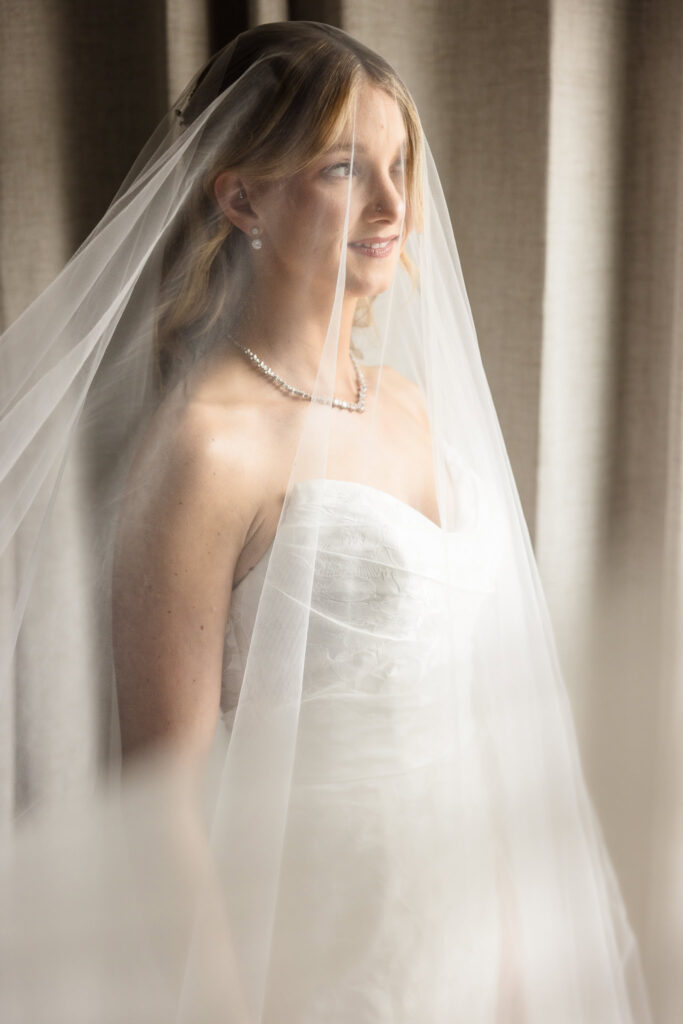 Bride standing by a window with her veil draped softly around her, glowing in natural light before the ceremony.