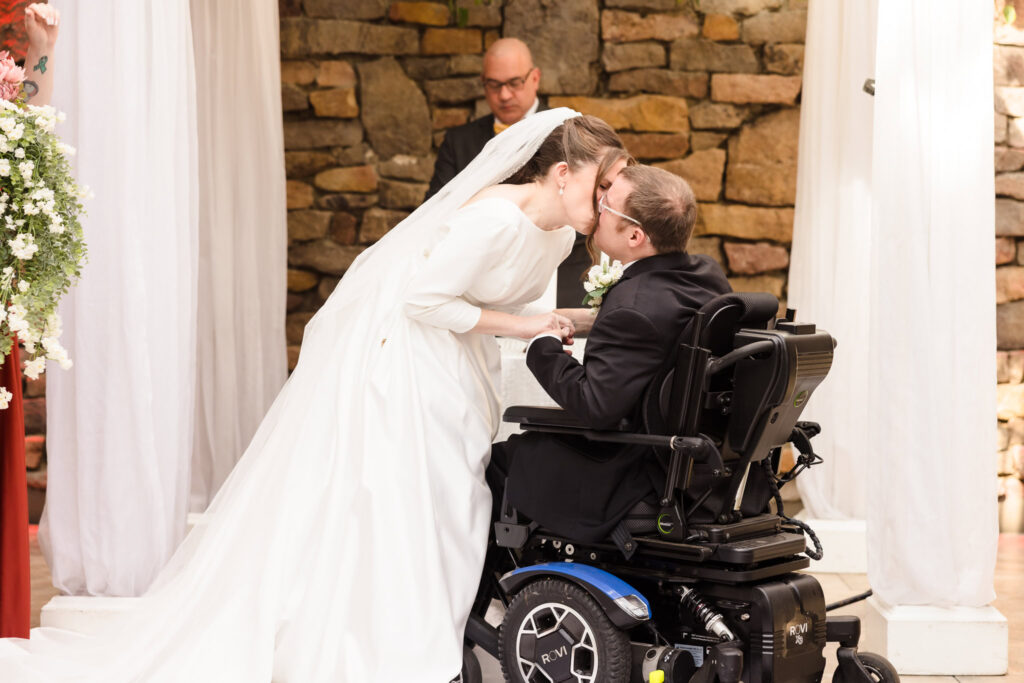 Bride leans in to kiss the groom during their heartfelt ceremony in front of a stone wall.