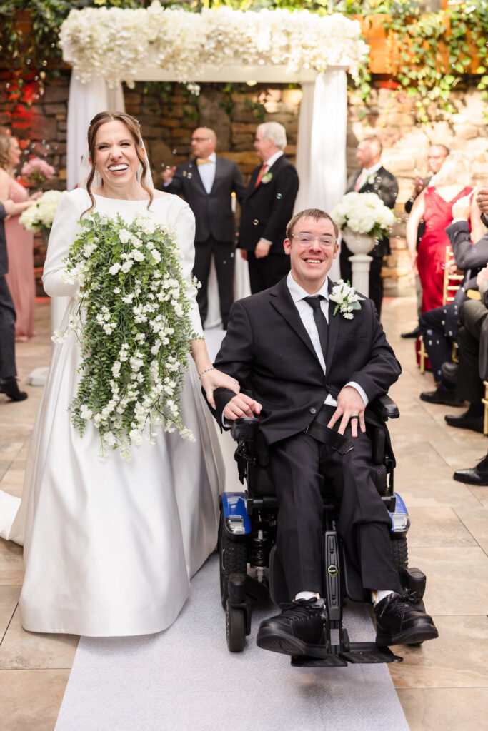 Bride and groom beam as they walk back down the aisle together under a white floral arch.
