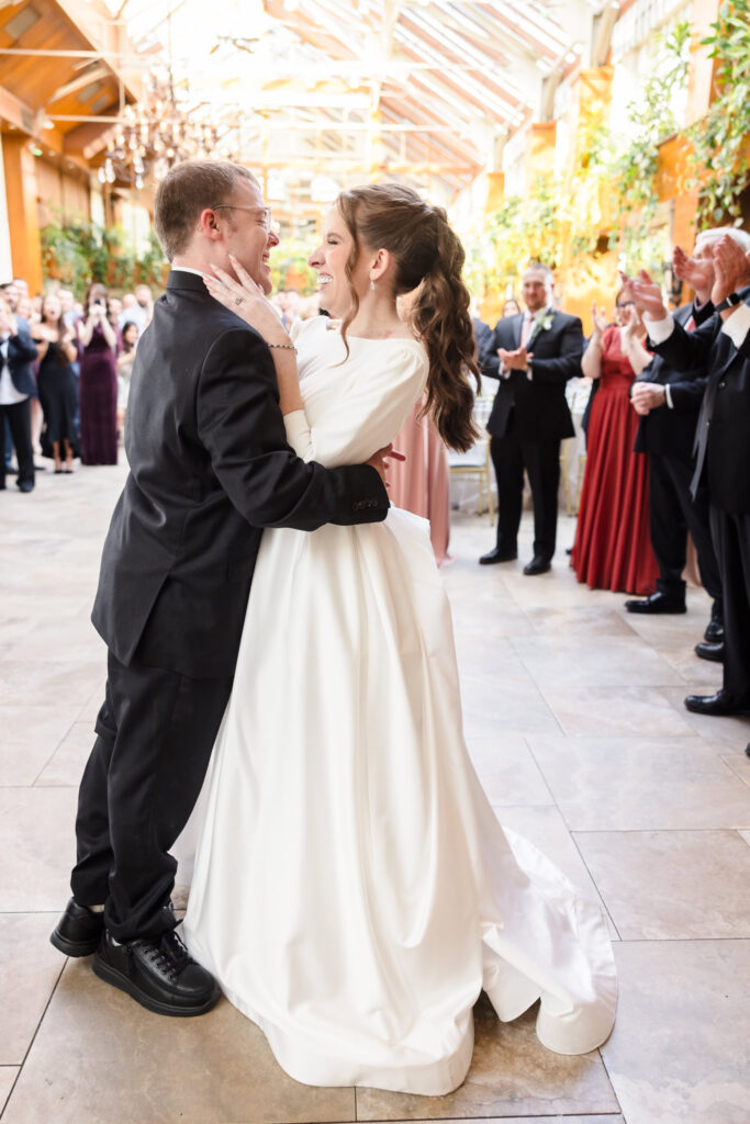 Bride and groom share their first dance in the Winter Garden surrounded by cheering friends and family.

