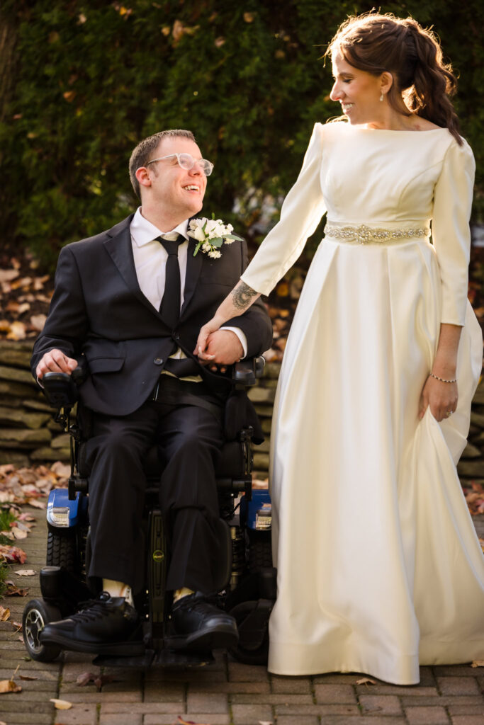 Bride holds groom’s hand while standing beside him on the landscaped paths at Fox Hollow, both smiling.