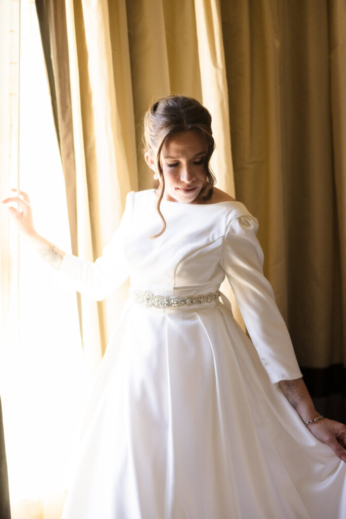 Bride looks down softly as she stands by a window in her wedding gown, illuminated by natural morning light.