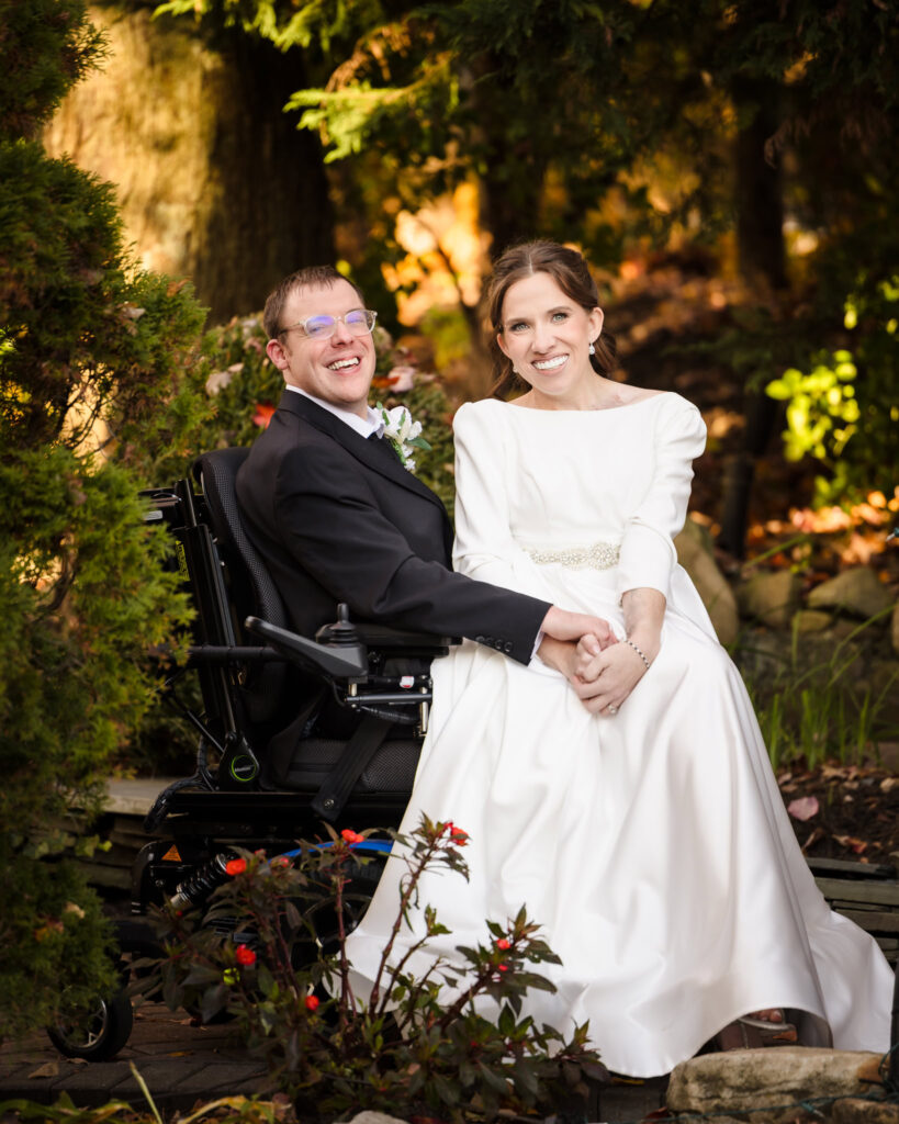 Bride and groom sit hand in hand in a serene garden moment framed by fall greenery.