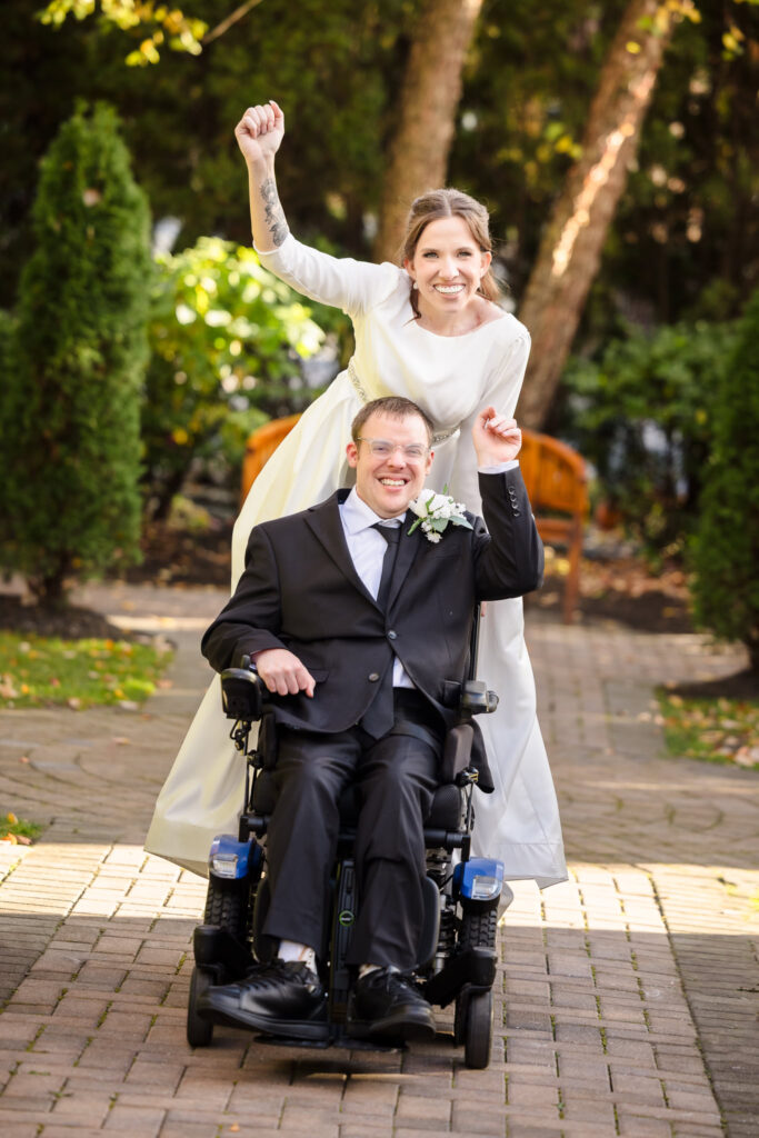 Bride raises her bouquet in celebration behind groom’s wheelchair as they smile together on a garden path.
