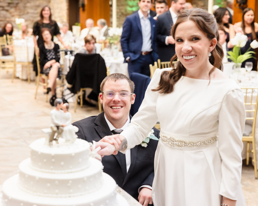 Bride and groom cut their white tiered wedding cake, smiling among guests during the reception.