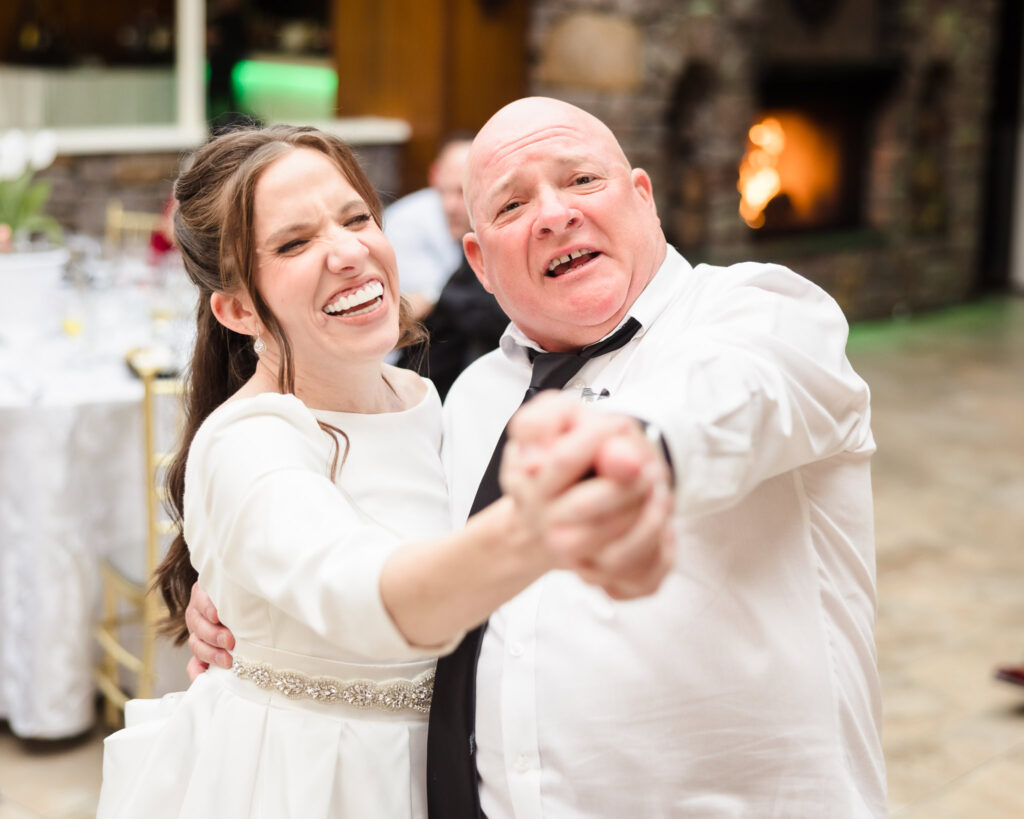 Bride laughs while dancing with her father, both holding hands with big smiles.