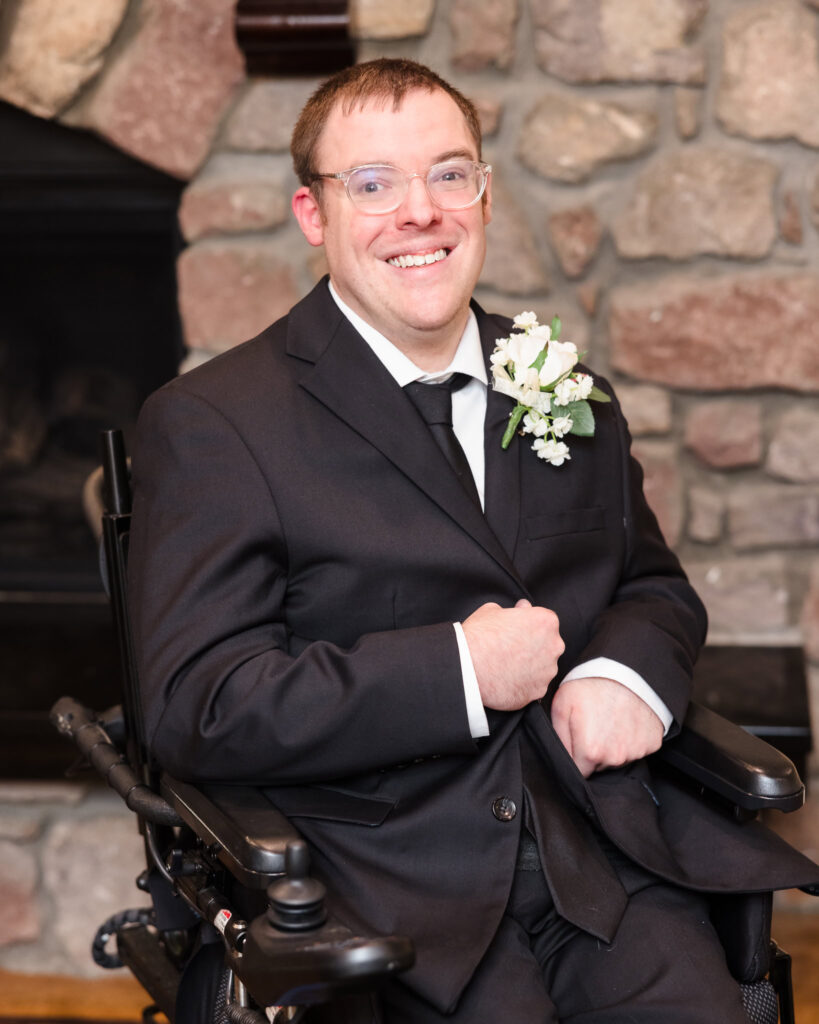 Groom smiles warmly while seated in his wheelchair, dressed in a black suit, in front of a stone fireplace.

